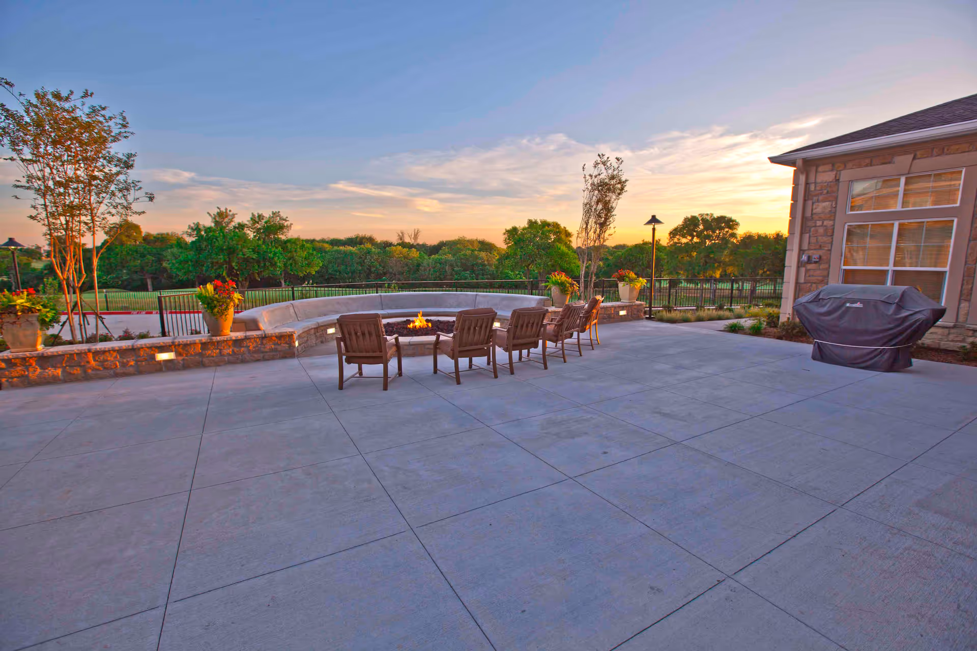 Outdoor patio area at sunset with a circular stone fire pit surrounded by wooden chairs and a curved stone bench. There are potted plants on the stone wall and a covered grill near the building with large windows. Trees and greenery are visible in the background.