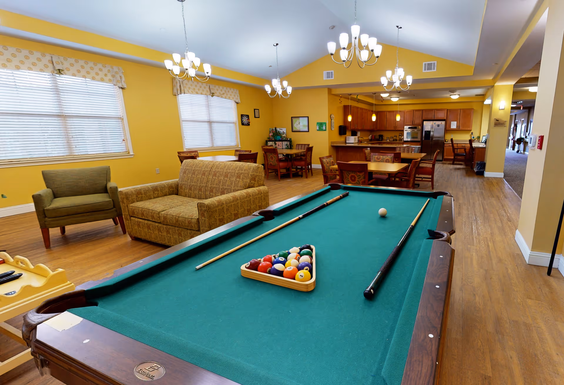 A spacious common area in an assisted living facility featuring a pool table with cues and balls set up, a green armchair and a patterned loveseat, multiple dining tables with chairs, and a kitchen area with wooden cabinets and stainless steel appliances. The walls are painted yellow, and there are large windows with blinds and valances. Several chandeliers hang from the ceiling providing light.