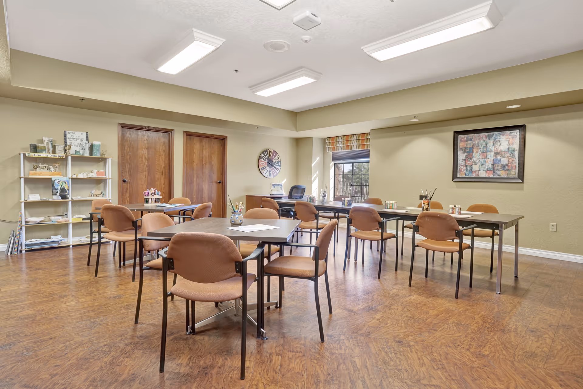 A bright community activity room with multiple tables and brown chairs on wood flooring, shelving, and wall art.