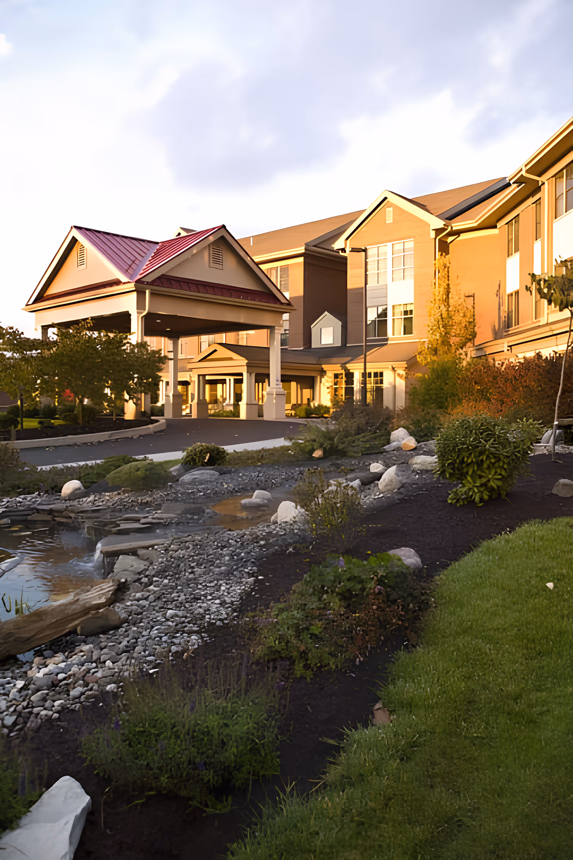 Exterior view of Bethany Village West facility during sunset, showing a large building with multiple windows and a covered entrance with a red roof. The foreground features a landscaped garden with rocks, shrubs, and a small water feature.