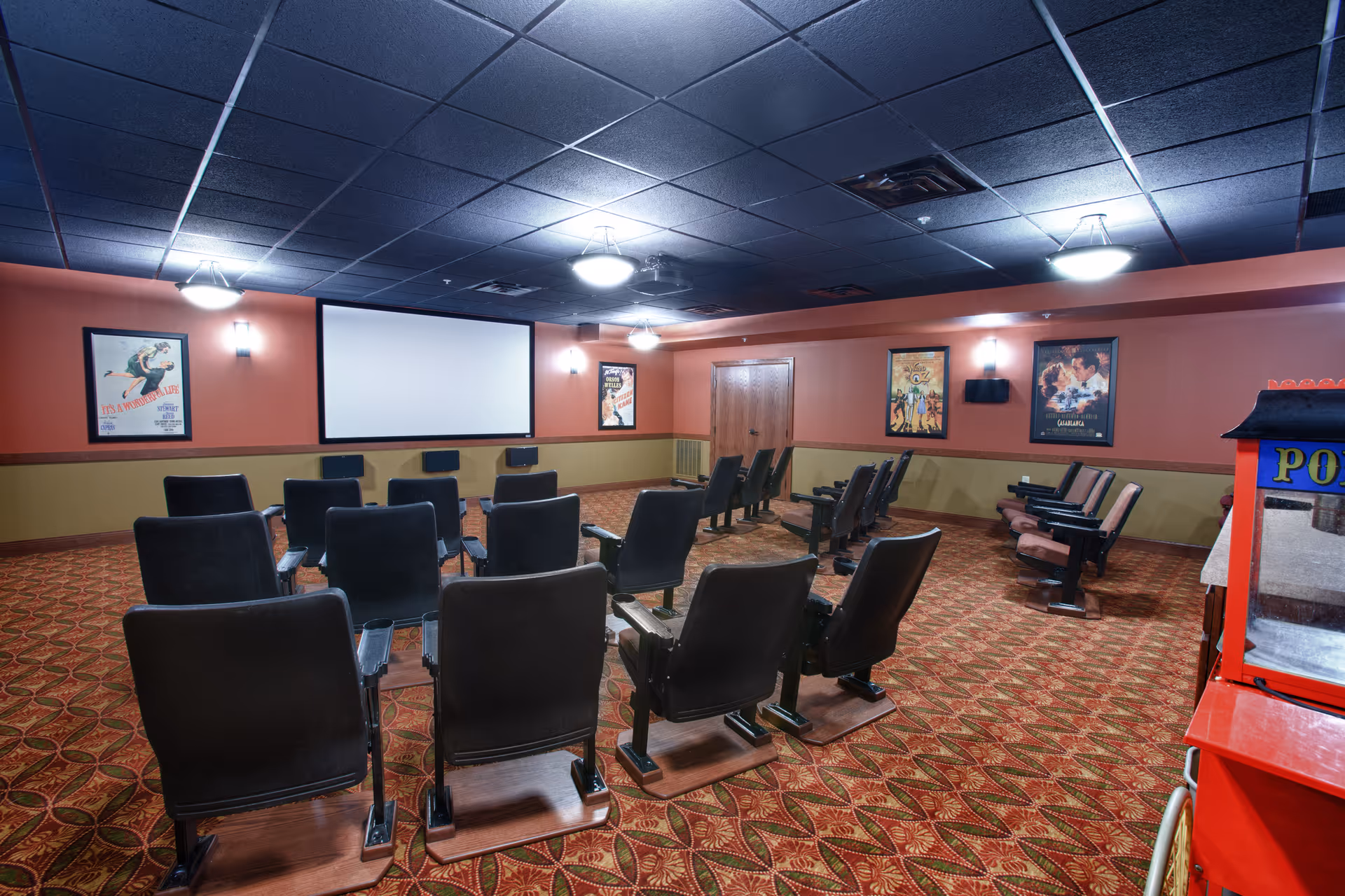 A small movie theater room with rows of black cushioned seats facing a large white projection screen. The walls are painted in two tones, with movie posters hanging on them. The carpet has a red and gold patterned design, and a popcorn machine is visible on the right side of the image.