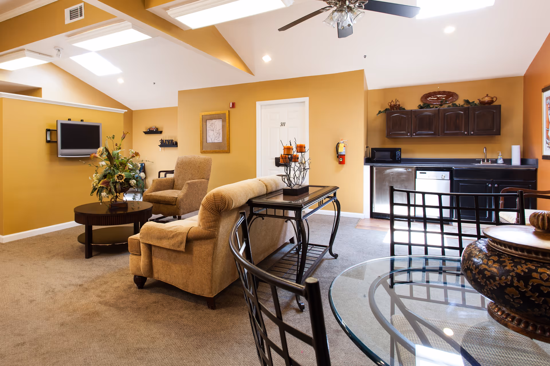 A cozy senior living common area with mustard yellow walls, beige carpet, and a vaulted ceiling with skylights. The room features a beige sofa and armchair, a round wooden coffee table with a floral arrangement, a wall-mounted flat-screen TV, and a small kitchenette with dark wood cabinets, a microwave, dishwasher, and sink. A glass dining table with black metal chairs is in the foreground.