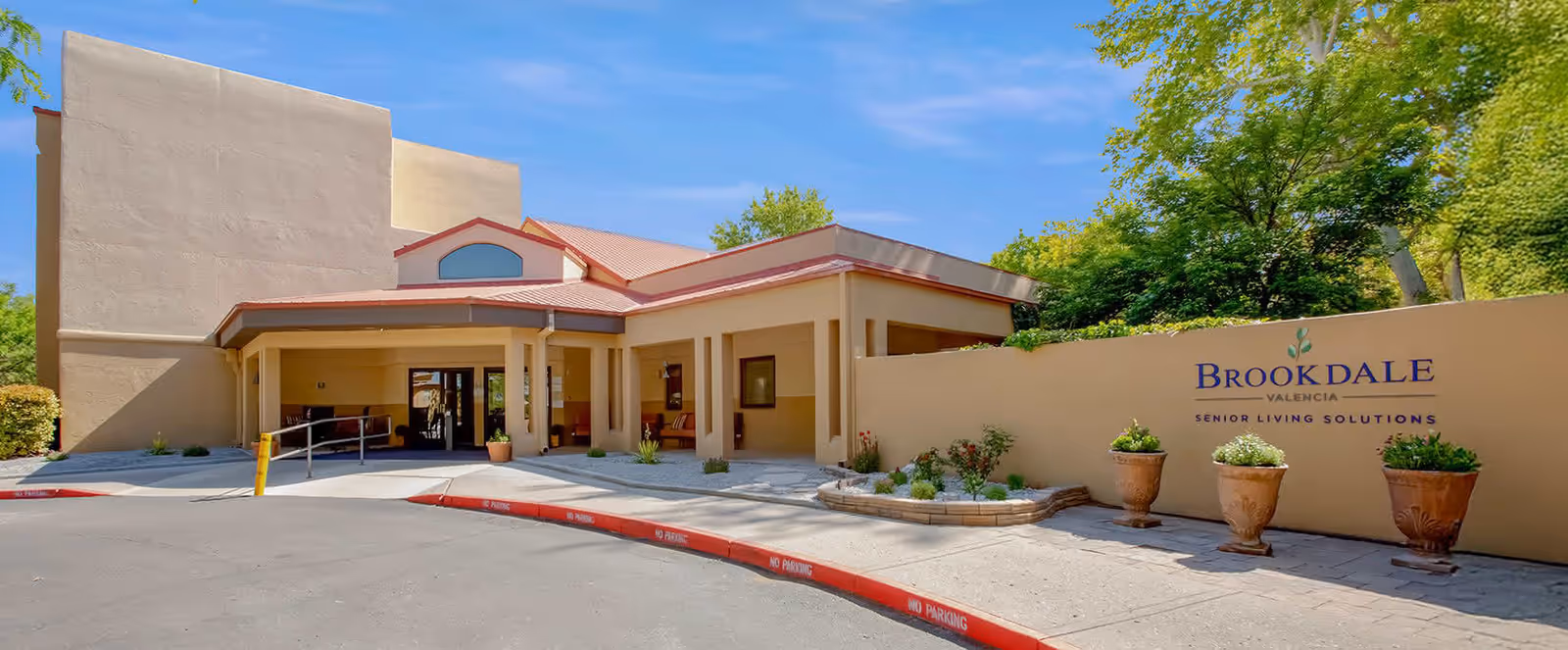 Exterior view of Brookdale Valencia senior living facility showing the entrance with a covered drop-off area, beige walls, and a sign on the right side that reads 'Brookdale Valencia Senior Living Solutions'. There are potted plants and greenery around the entrance under a clear blue sky.