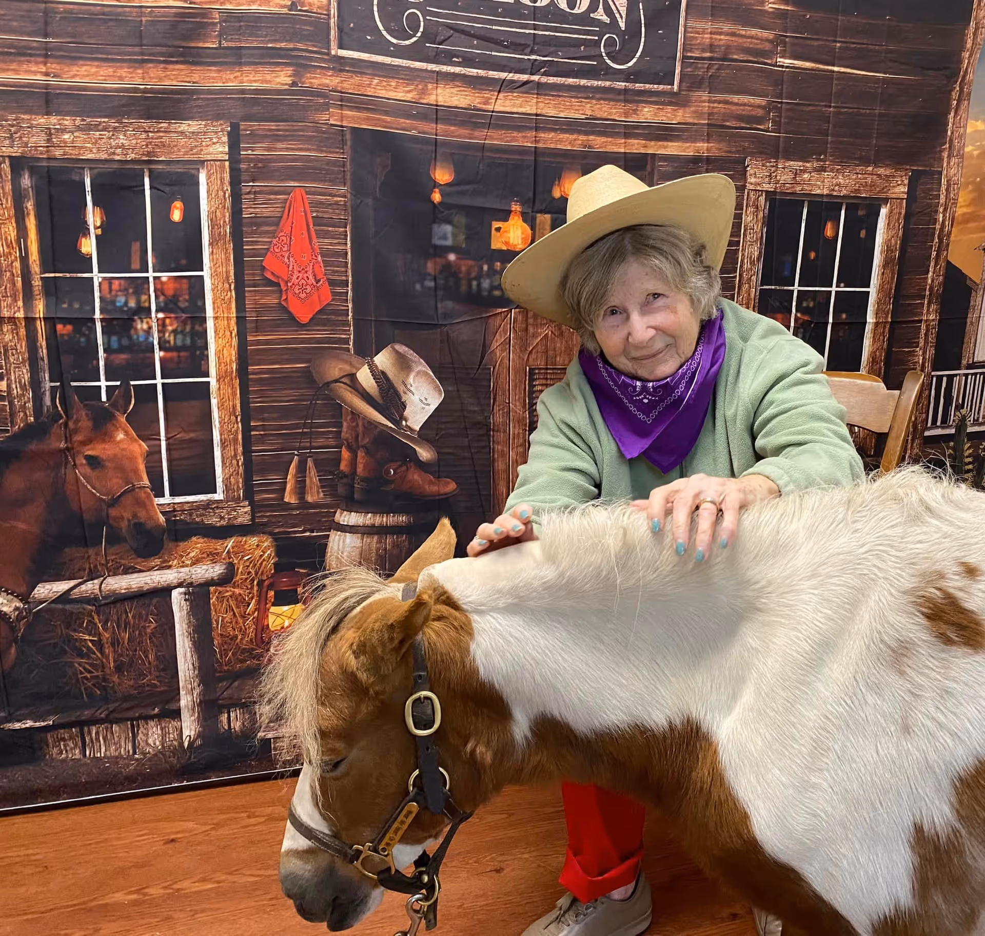 An elderly woman wearing a cowboy hat and purple bandana smiles while petting a small brown and white pony indoors. The background features a western-themed backdrop with a wooden building facade, windows, a horse, cowboy boots, and a red bandana hanging on the wall.