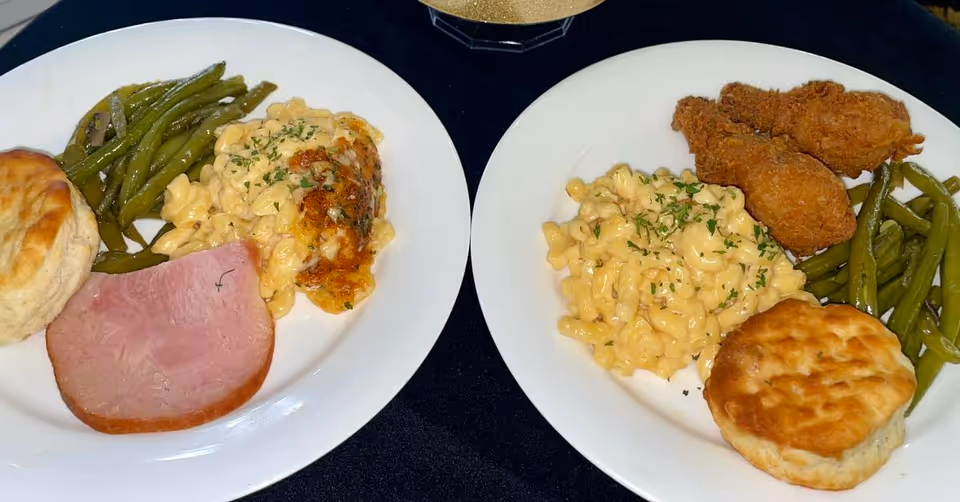 Two white plates of food on a black surface. One plate has green beans, macaroni and cheese, a biscuit, and a slice of ham. The other plate has green beans, macaroni and cheese, a biscuit, and two pieces of fried chicken.
