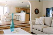 A bright and spacious living room area with a beige sofa adorned with patterned pillows. In the foreground, there is a white coffee table with a tall blue glass vase and decorative items. The living room opens up to a dining area with a white dining table and chairs, and a kitchen with wooden cabinets and a breakfast bar with stools. The space features wooden flooring and an archway separating the living room from the kitchen and dining area.