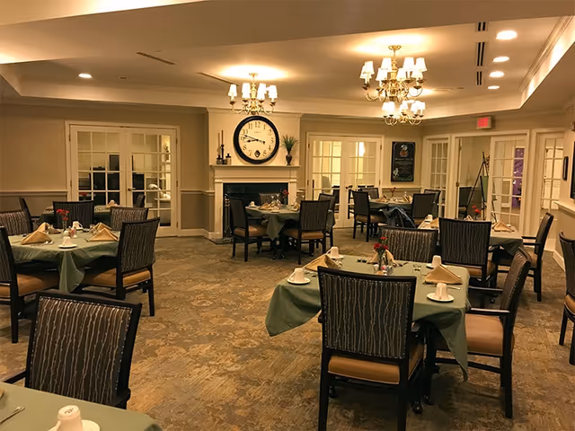 A dining room with multiple tables covered in green tablecloths, each set with napkins, cups, and small flower arrangements. The room features a large clock above a fireplace, chandeliers hanging from the ceiling, and glass-paneled doors along the walls.