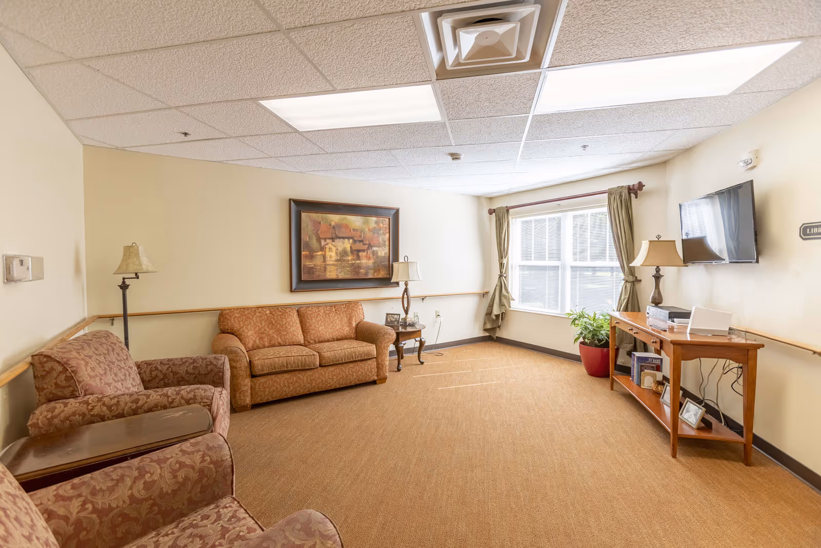 A cozy living room area with two patterned armchairs and a matching sofa, a wooden side table with a lamp, a framed painting on the wall, a large window with green curtains, a wooden console table with a lamp, books, and a flat-screen TV mounted on the wall.