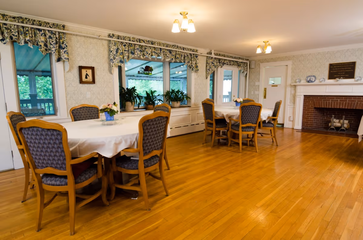 A dining room with two round tables covered with white tablecloths, each surrounded by six wooden chairs with blue patterned upholstery. The room has hardwood floors, floral valances over the windows, several potted plants on the windowsill, and a brick fireplace with a white mantel on the right side. The walls are decorated with light-colored wallpaper and a framed picture and plaque are visible.