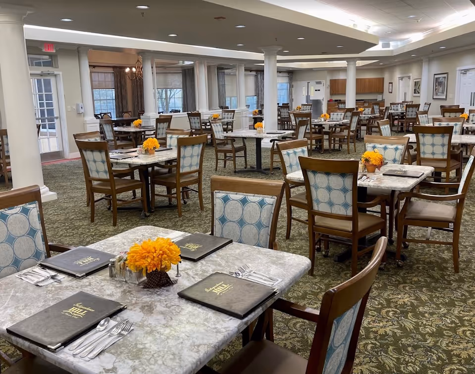 A spacious dining room with multiple tables and chairs arranged neatly. Each table has a small orange flower centerpiece and menus placed on top. The room features carpeted floors with a floral pattern, white columns, and large windows with curtains. The lighting is bright with ceiling lights, and there is a kitchen area visible in the background.