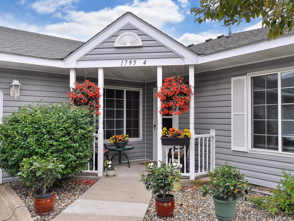 Entrance of a gray single-story building with white trim, featuring a small covered porch with two hanging red flower baskets and several potted plants on the ground and porch. The building number 1795 4 is displayed above the porch. There is a window with white shutters on the right side and a small round table with chairs on the porch.