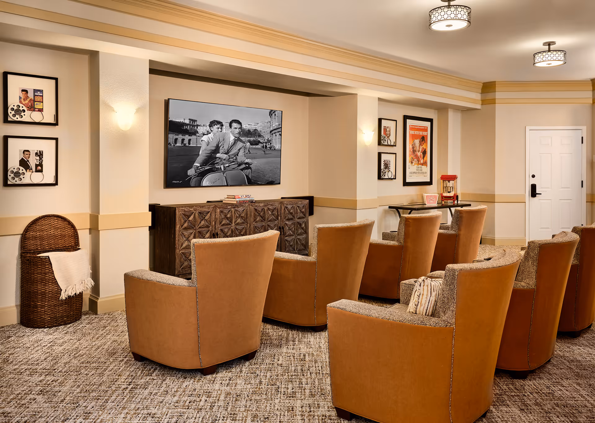 A cozy movie room with several brown upholstered chairs arranged in rows facing a wall-mounted TV displaying a black and white movie scene. The room features warm lighting, framed vintage movie posters on the walls, a decorative wooden cabinet beneath the TV, a popcorn machine on a small table, and a woven basket with a blanket in the corner.