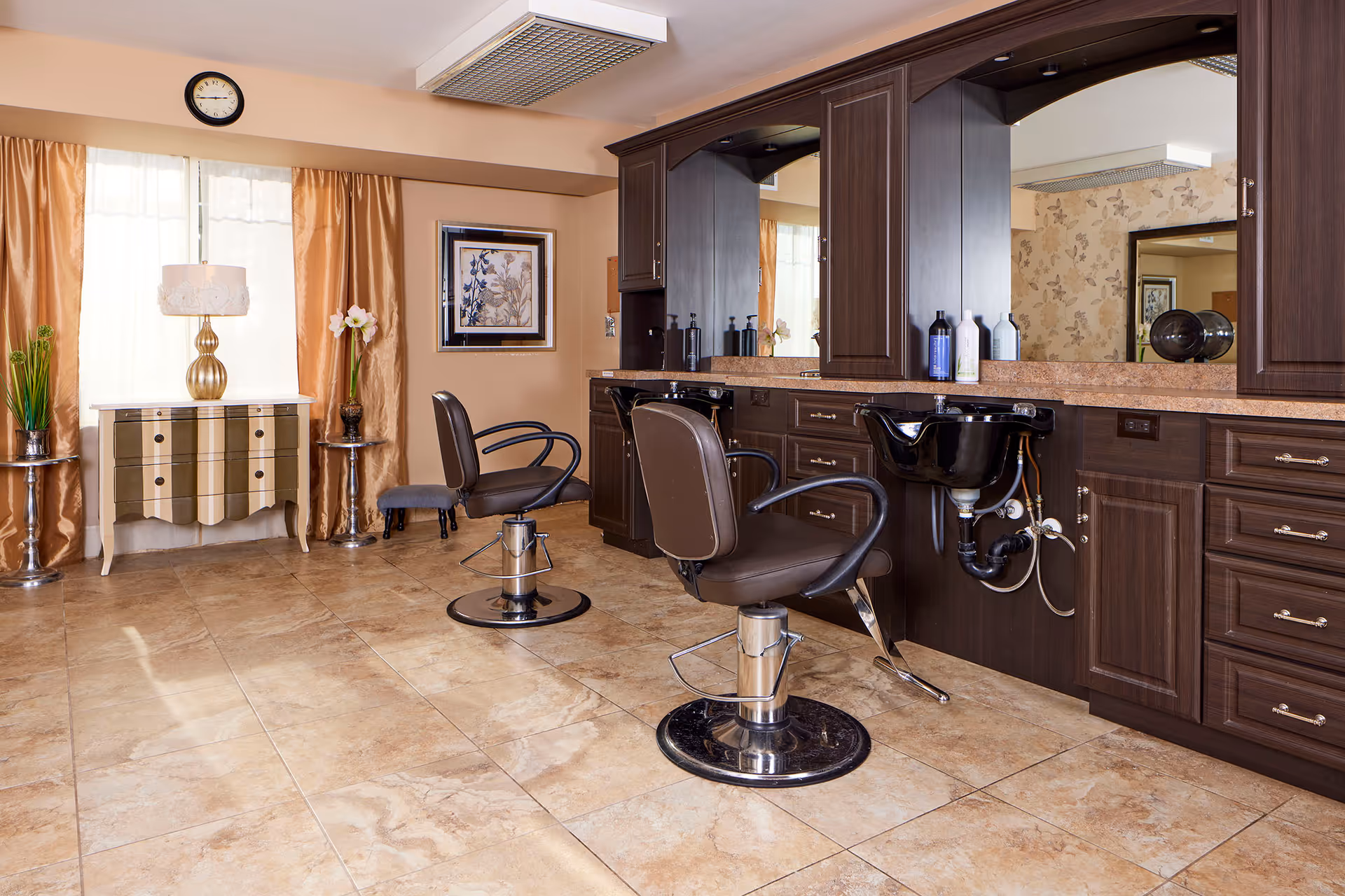 Interior of a senior living facility's hair salon with two brown salon chairs in front of a large mirror and dark wood cabinetry. There is a black hair washing sink attached to the cabinetry. The room has beige tiled floors, a small decorative table with a lamp and flowers near a window with gold curtains, and a wall clock above the window.