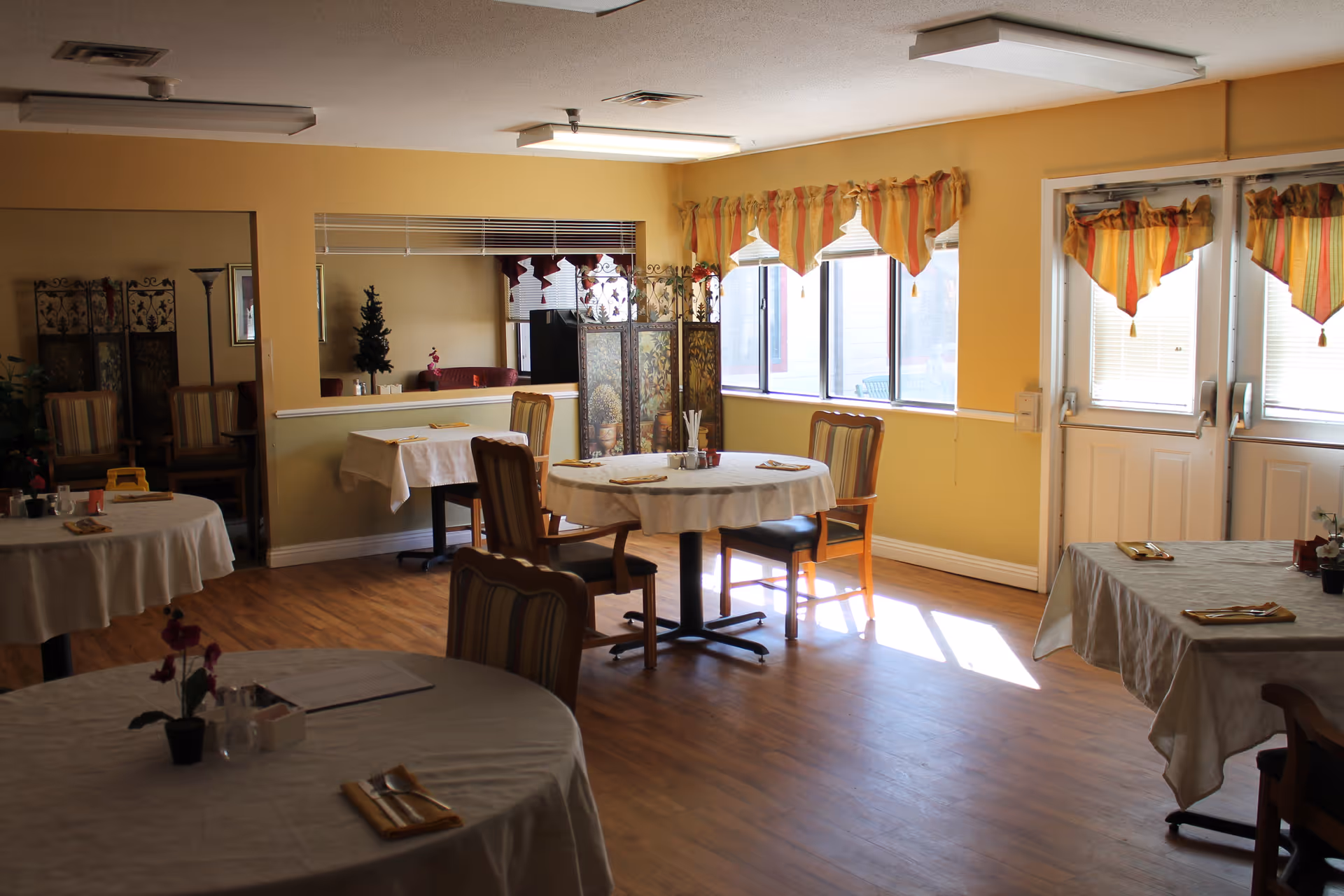 A bright dining room with several round and rectangular tables covered with white tablecloths, each set with napkins and utensils. The room has wooden flooring, yellow walls, large windows with striped valances, and double doors letting in natural light. Chairs with striped upholstery are arranged around the tables, and a decorative folding screen and small plants are visible in the background.