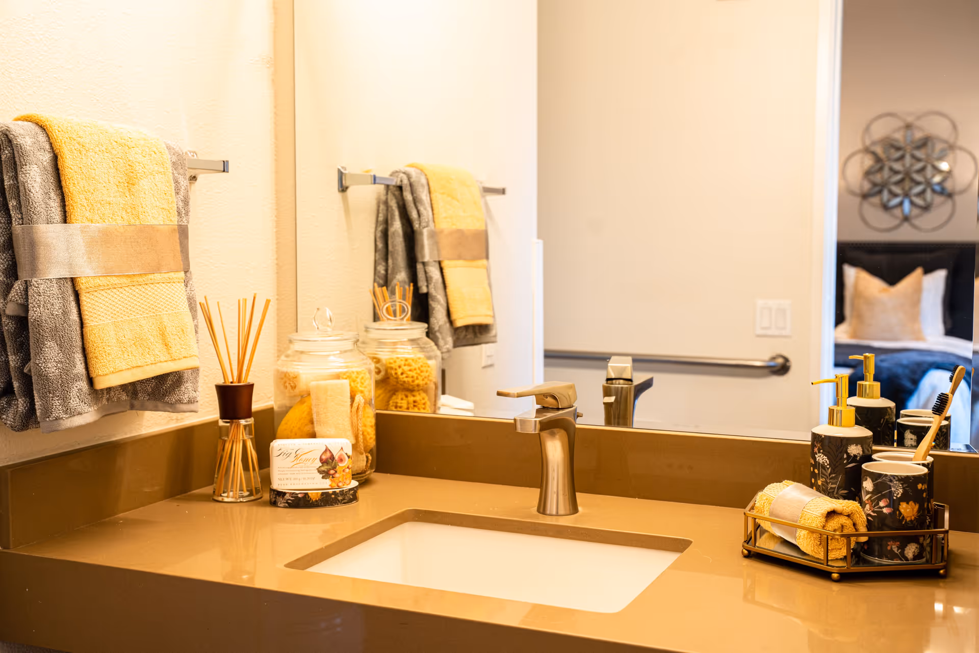 A bathroom sink countertop with a modern faucet, decorated with neatly folded towels, a glass jar filled with bath sponges, a reed diffuser, and a tray holding soap dispensers and a toothbrush holder. A large mirror reflects part of a bedroom with a bed and decorative wall art.