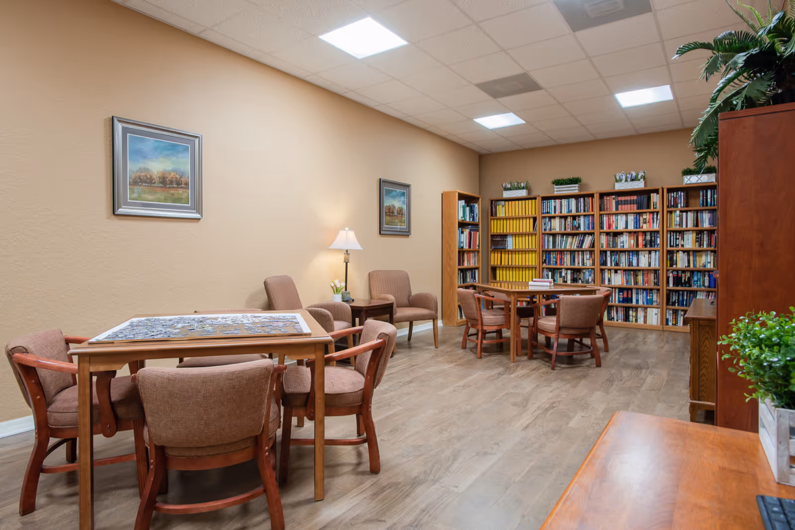 A cozy reading and activity room with wooden tables and upholstered chairs arranged for group activities. One table has a partially completed puzzle. The room features bookshelves filled with books and decorative plants on top. The walls are painted beige and adorned with framed landscape paintings. The floor is wood, and the ceiling has recessed lighting.