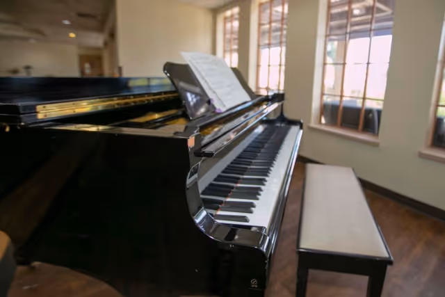A black grand piano with sheet music on the music stand, positioned in a room with large windows letting in natural light. A piano bench is placed next to the piano on a wooden floor.