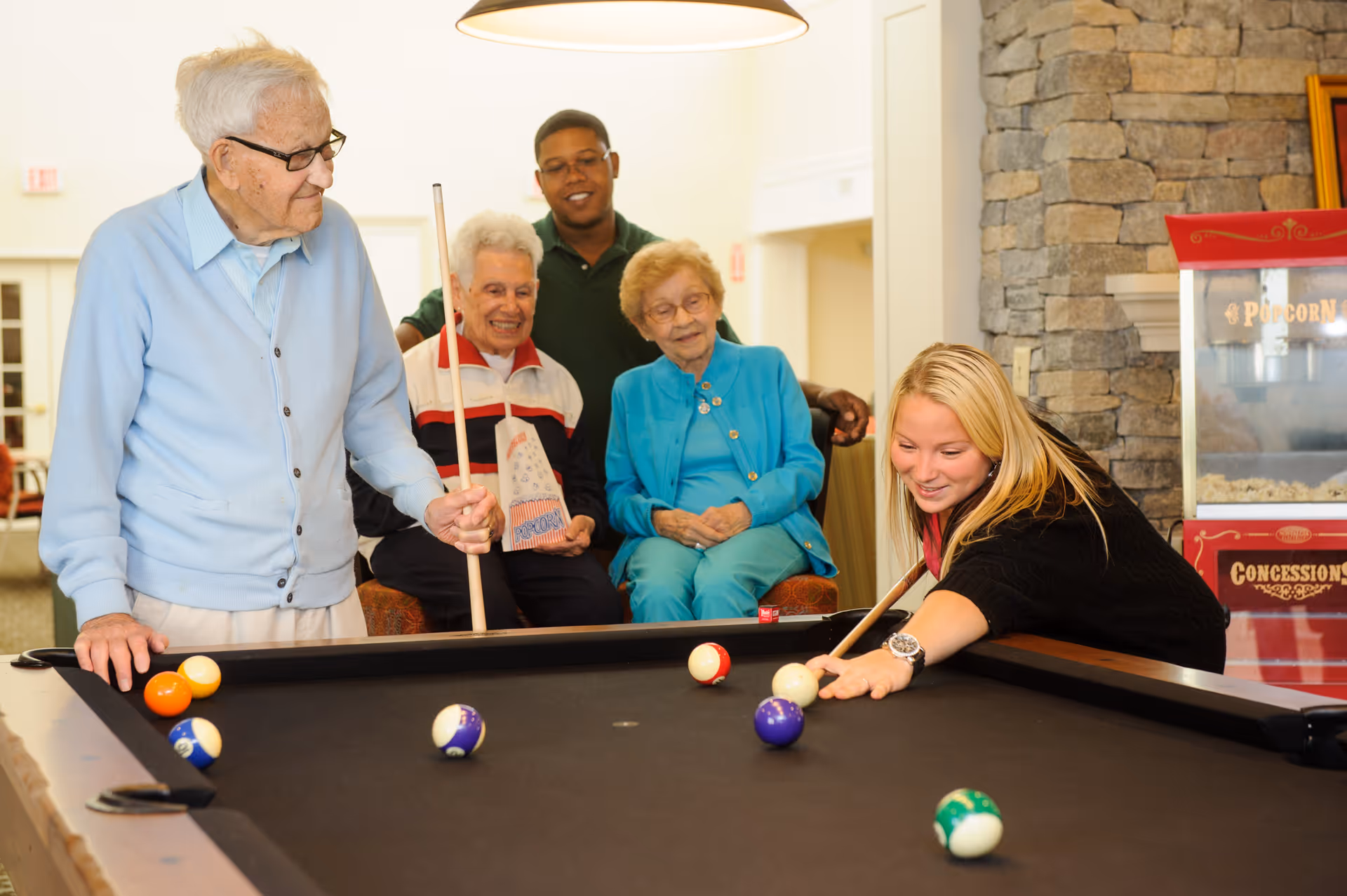 A group of elderly people and a young woman playing pool in a recreational room. The young woman is taking a shot while the elderly man next to her holds a pool cue. Two elderly women and a young man watch and smile in the background. The room has a stone wall with a popcorn machine and a warm, inviting atmosphere.