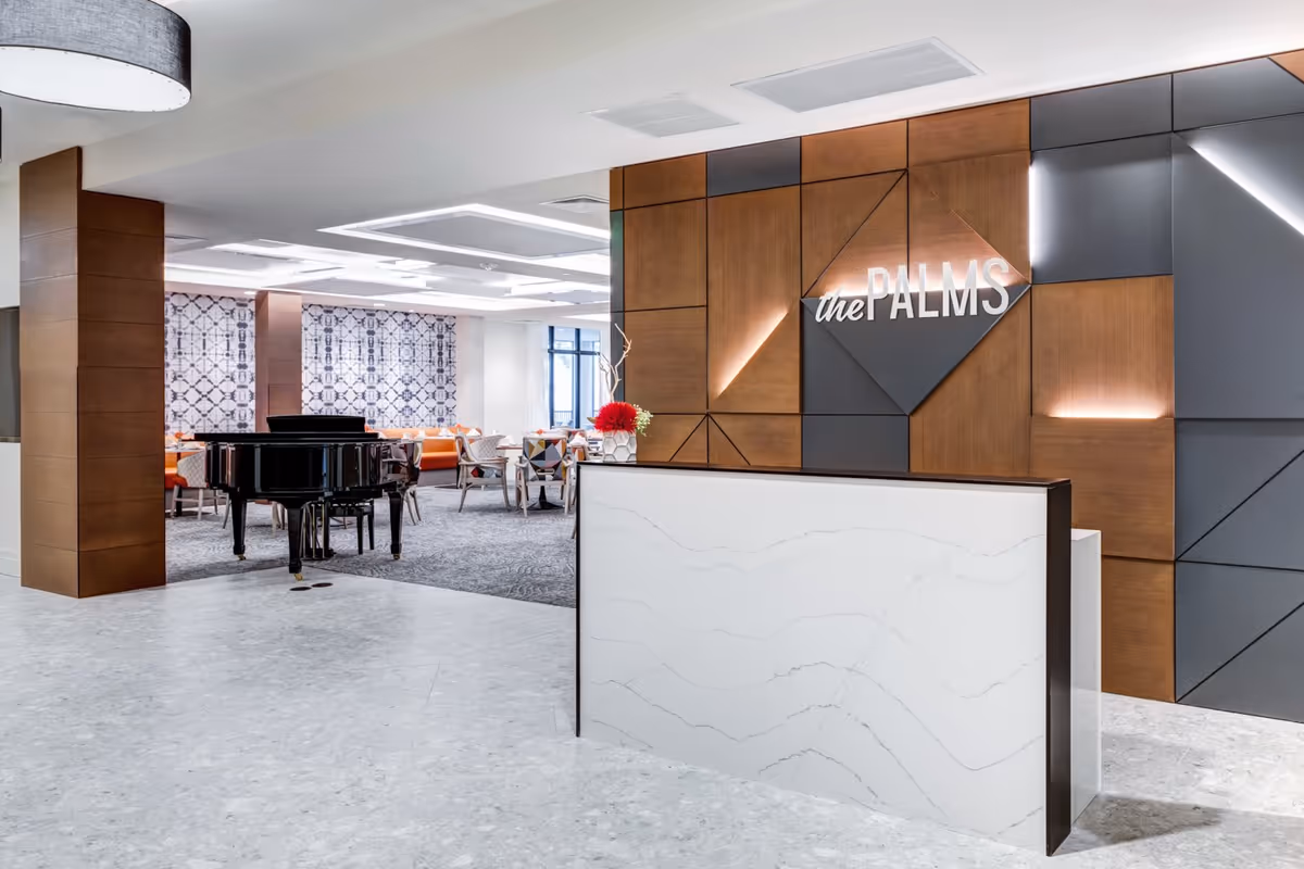 Modern reception area with a white marble desk in the foreground and a decorative wall behind it featuring the illuminated sign 'the PALMS'. In the background, there is a black grand piano and a dining area with tables and chairs, some with orange cushions, and patterned curtains on the windows.