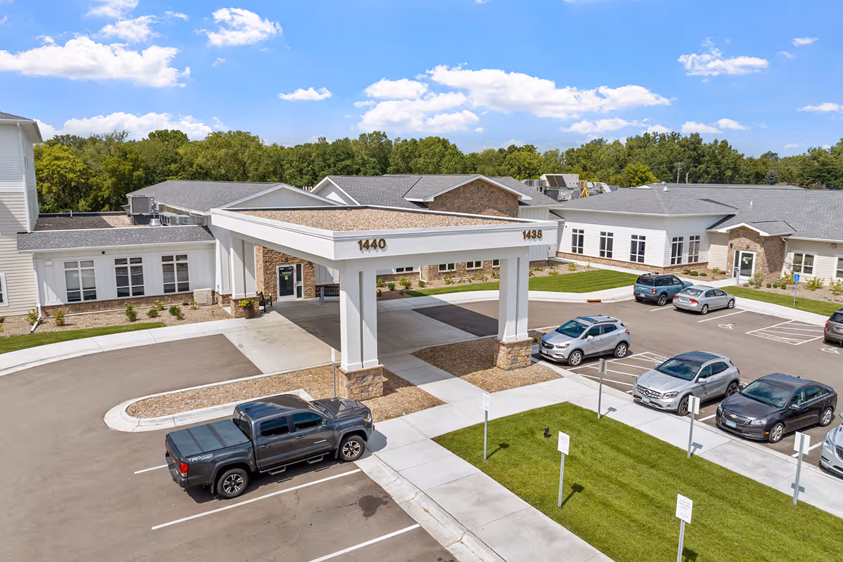 Front entrance and porte-cochère of a senior living facility with a parking lot, several cars, and surrounding buildings.