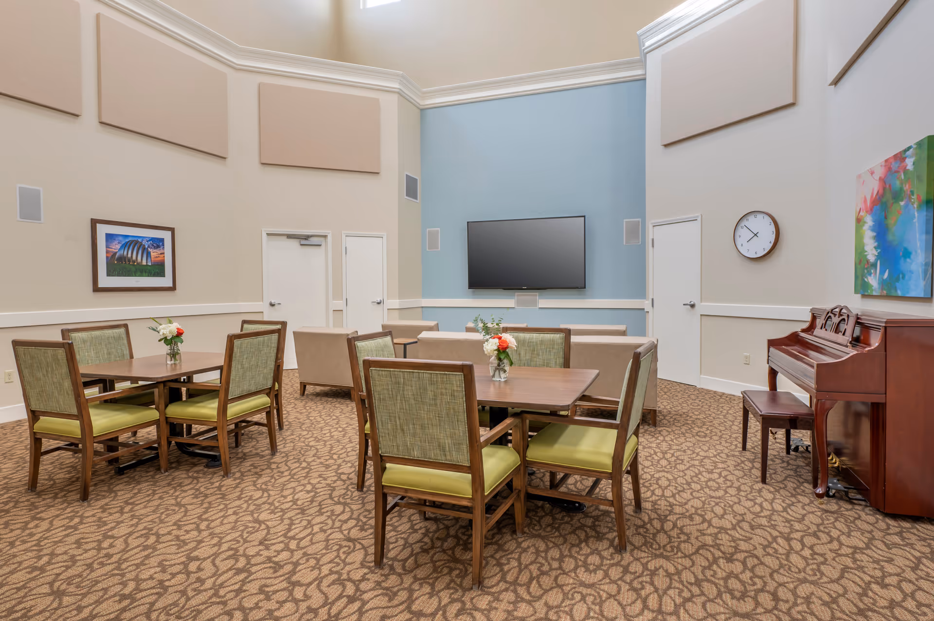 A bright communal room with tables and green-upholstered chairs, a wall-mounted TV, sofa seating and a piano.