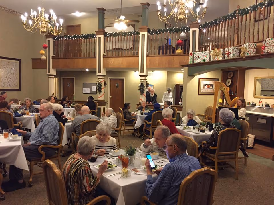 A dining room in a senior living facility decorated for the holidays with garlands and wrapped presents on the balcony railing. Several elderly residents are seated at tables covered with white tablecloths, eating and socializing. A woman is playing a harp in the corner of the room.