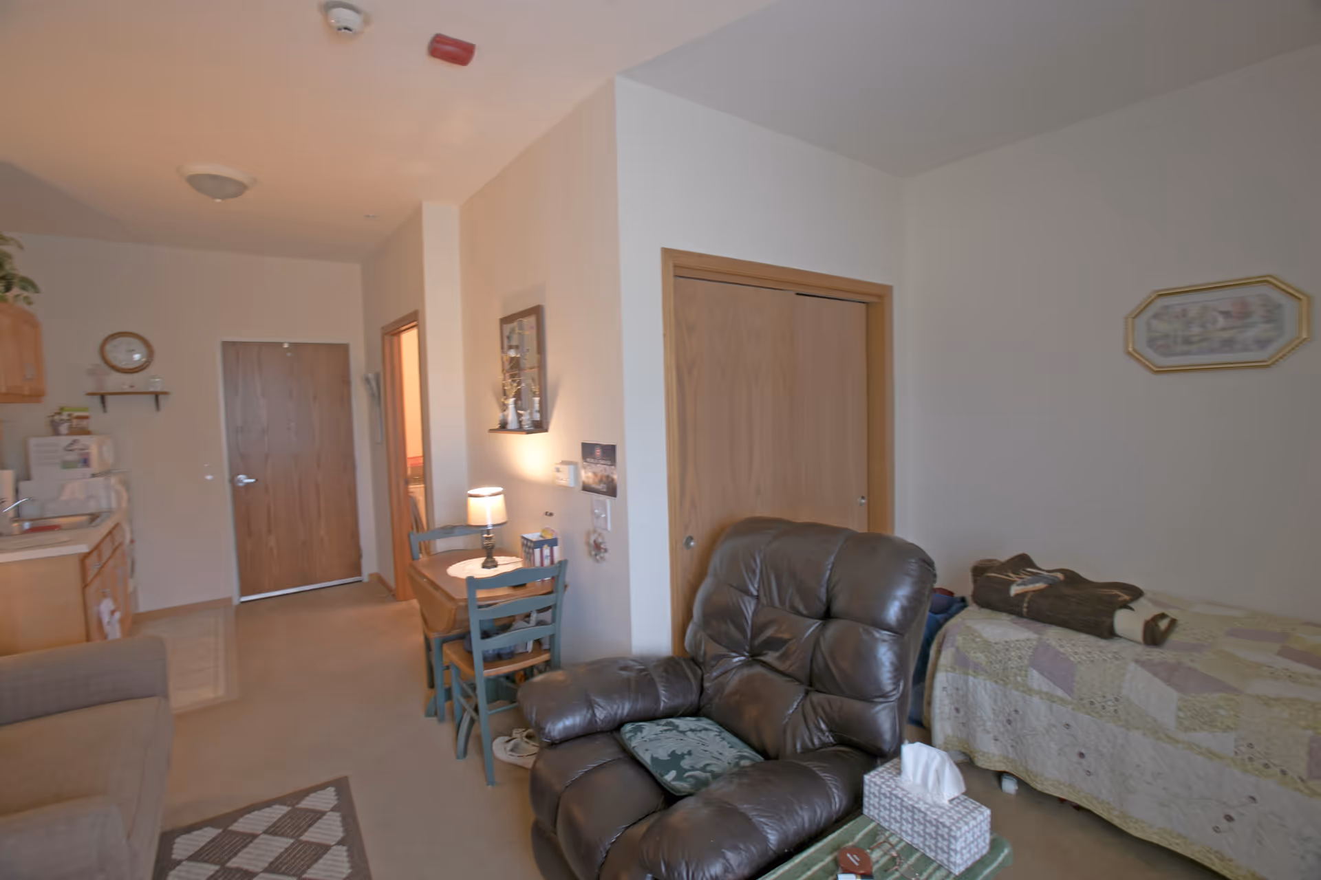 Interior view of a senior living facility room at Heritage Woods of Yorkville showing a cozy living area with a dark brown recliner chair, a small table with a tissue box, a single bed with a patchwork quilt, a small dining table with two chairs and a lamp, and a kitchenette area with cabinets, a microwave, and a sink. The room has beige walls and carpeted floor.