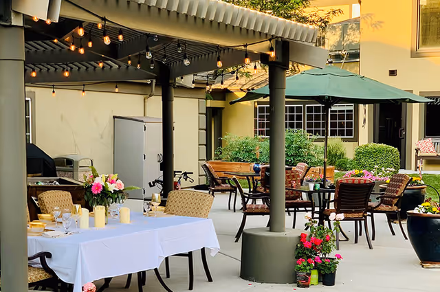 Outdoor patio area at Cedarwood at Sandy with tables and chairs arranged for dining and relaxation. One table is covered with a white tablecloth and decorated with flowers and candles. String lights hang from the pergola overhead, and there are potted plants and greenery around the space.