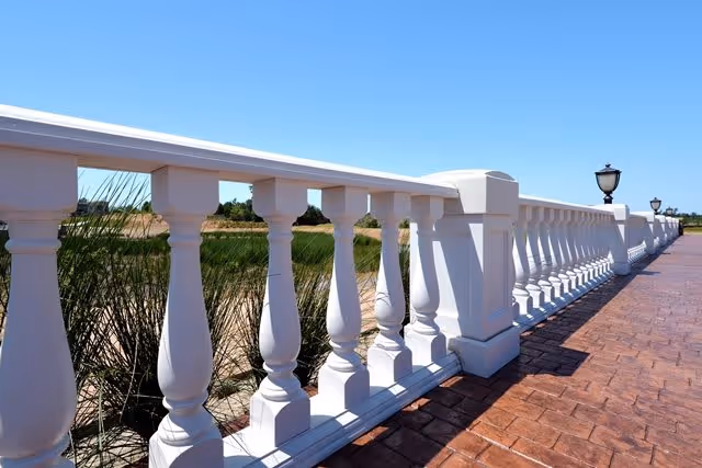 A white balustrade railing along a paved walkway with decorative street lamps, overlooking a natural area with grass and water under a clear blue sky.