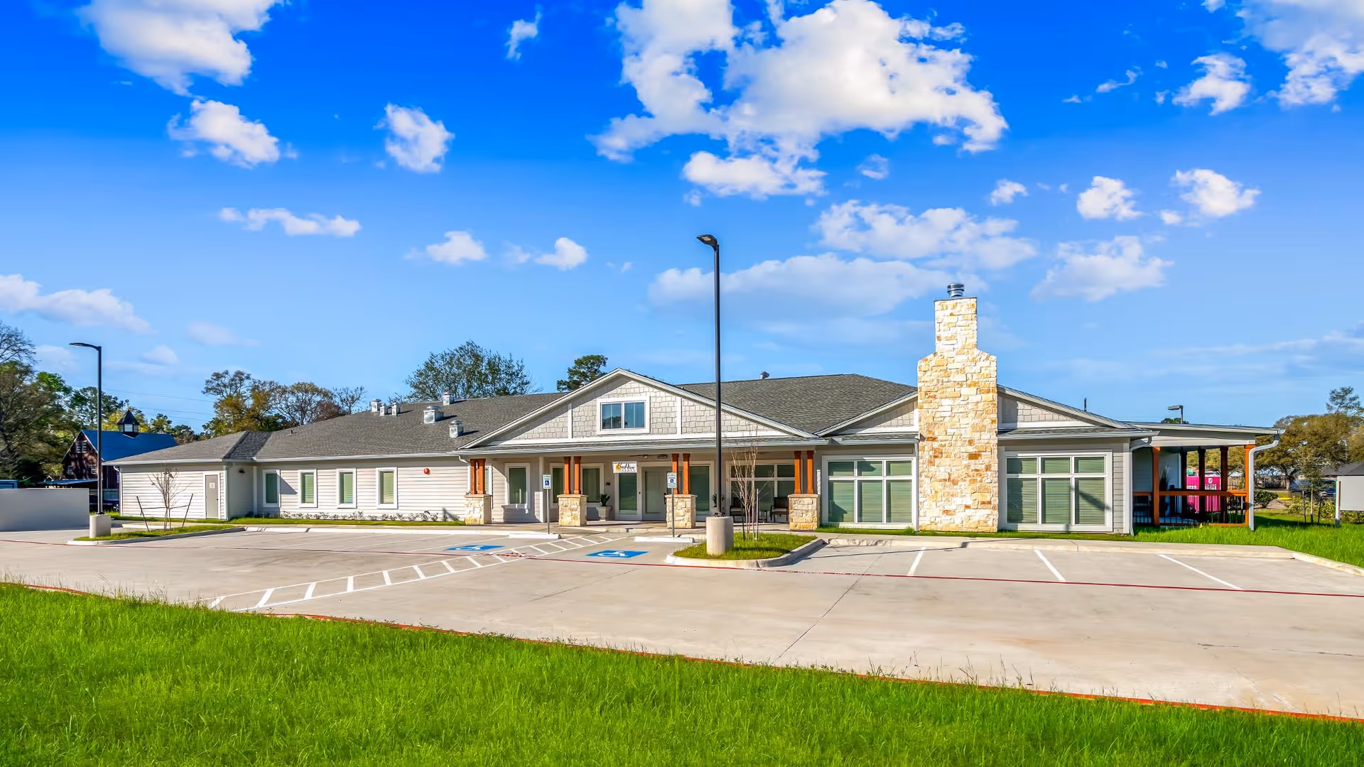 Exterior view of a single-story senior living facility building with a stone chimney, multiple windows, and a covered entrance. The building is surrounded by a parking lot with marked spaces and green grass in the foreground under a blue sky with scattered clouds.