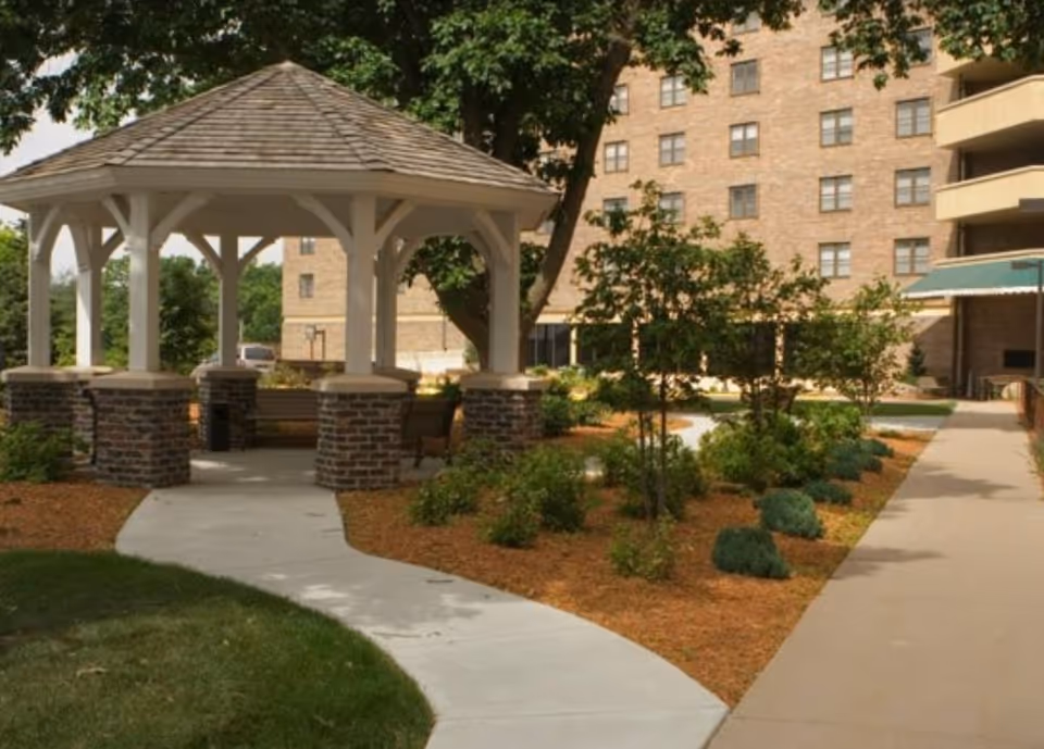 Outdoor gazebo with a shingled roof and brick pillars surrounded by landscaped garden beds and trees, with a multi-story brick building in the background.