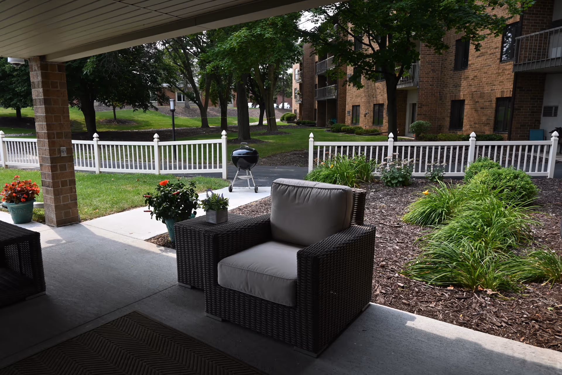 Covered patio with a cushioned wicker armchair and side table facing landscaped grounds, a white fence, and a brick building.