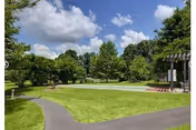 A well-maintained outdoor area with a paved walking path winding through green grass and trees under a partly cloudy blue sky. There is a wooden pergola structure on the right side and benches along the path.