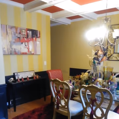 A dining room with a glass-top table surrounded by six wooden chairs with white cushions. The table is decorated with various colorful ornaments and a centerpiece. The walls are painted with yellow vertical stripes and solid yellow. A modern abstract painting hangs on one wall. A chandelier with multiple lights hangs above the table. There is a black sideboard against the wall with small decorative items on it.