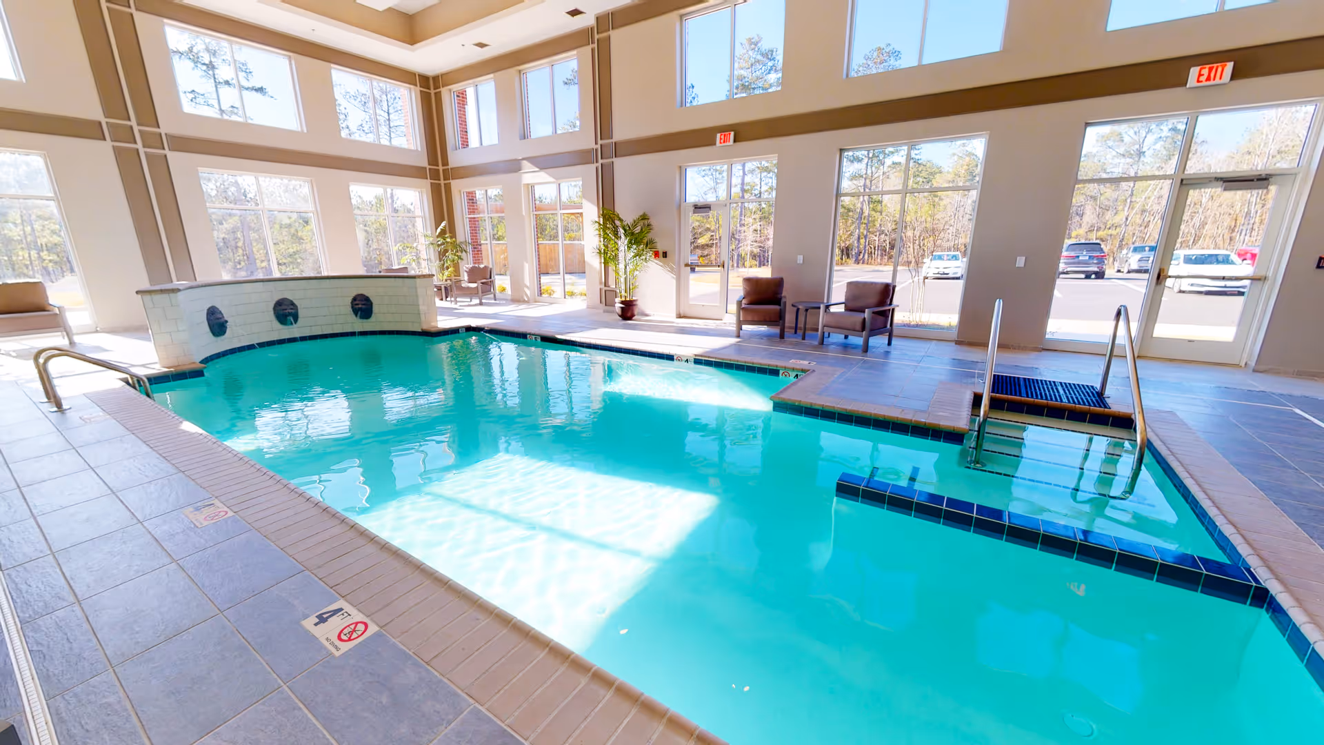 Indoor swimming pool with clear blue water in a bright room with large windows allowing natural light. There are chairs and small tables near the windows, and a door leading outside. The pool area has tiled flooring and metal handrails for entering the pool.