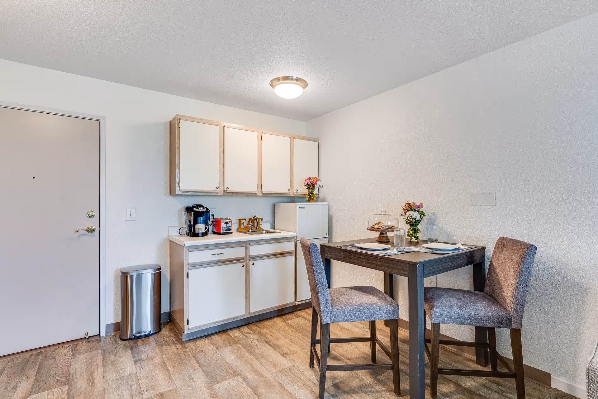 A small kitchen area with white cabinets, a countertop holding a coffee maker, toaster, and decorative letters spelling 'EAT'. Next to the kitchen is a small dining table set for two with placemats, glasses, and a cake stand with pastries. Two gray upholstered chairs are placed at the table. The floor has wood-style flooring, and the walls are plain white. A closed door and a stainless steel trash can are visible on the left side.
