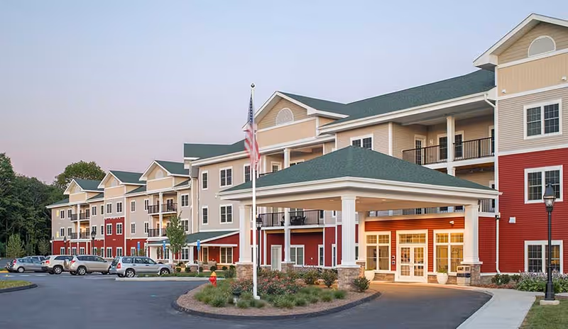 Exterior view of a multi-story senior living facility building with beige and red siding, green roofs, balconies, and a covered entrance. Several cars are parked in front, and there is an American flag on a flagpole near the entrance.