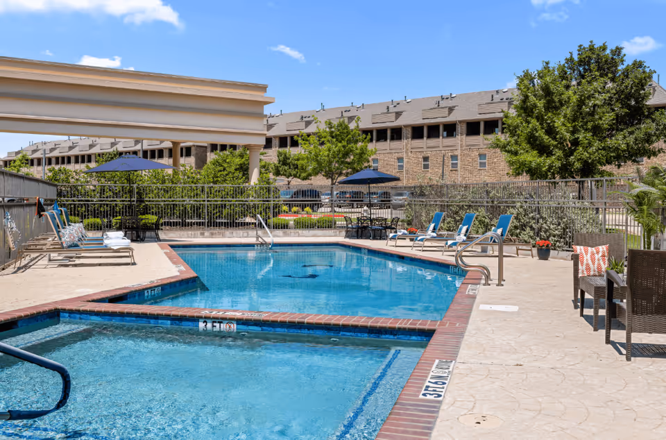 Outdoor swimming pool and lounge area with chairs, umbrellas, and apartment buildings in the background.