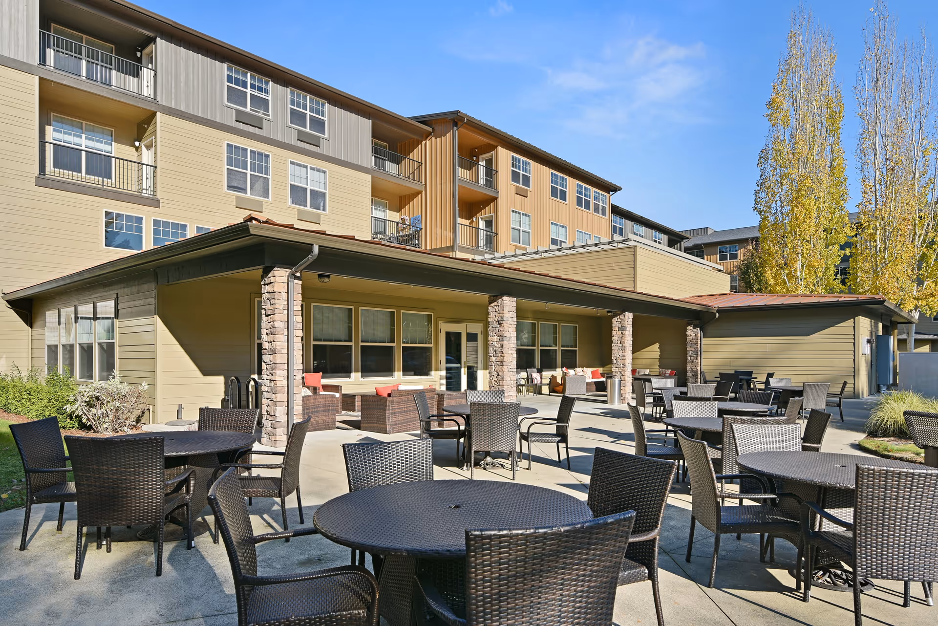 Outdoor patio area at Bonaventure of Lacey with multiple round tables and wicker chairs arranged on a concrete surface. The patio is adjacent to a building with stone pillars and large windows. Trees with autumn foliage are visible in the background under a clear blue sky.