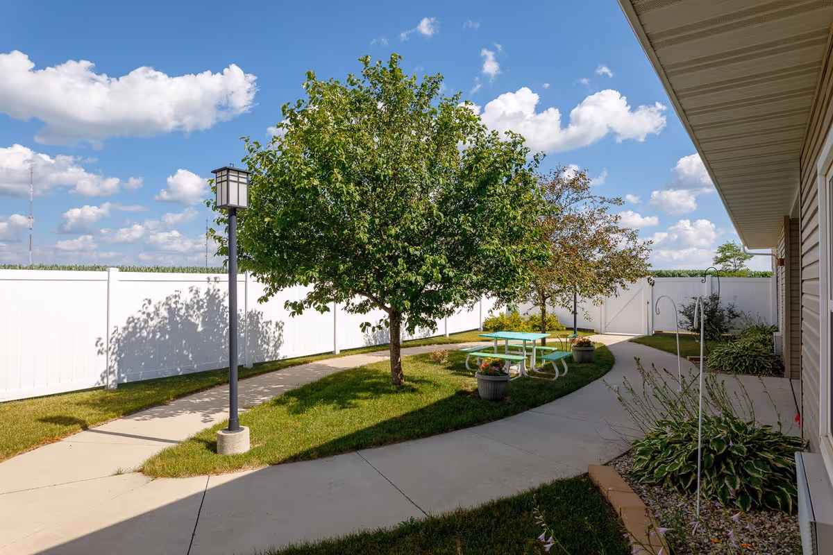 Outdoor courtyard area with a concrete walkway curving around a grassy section with two trees, a green picnic table with benches, potted plants, and a lamp post. The area is enclosed by a white fence and the sky is blue with scattered clouds.