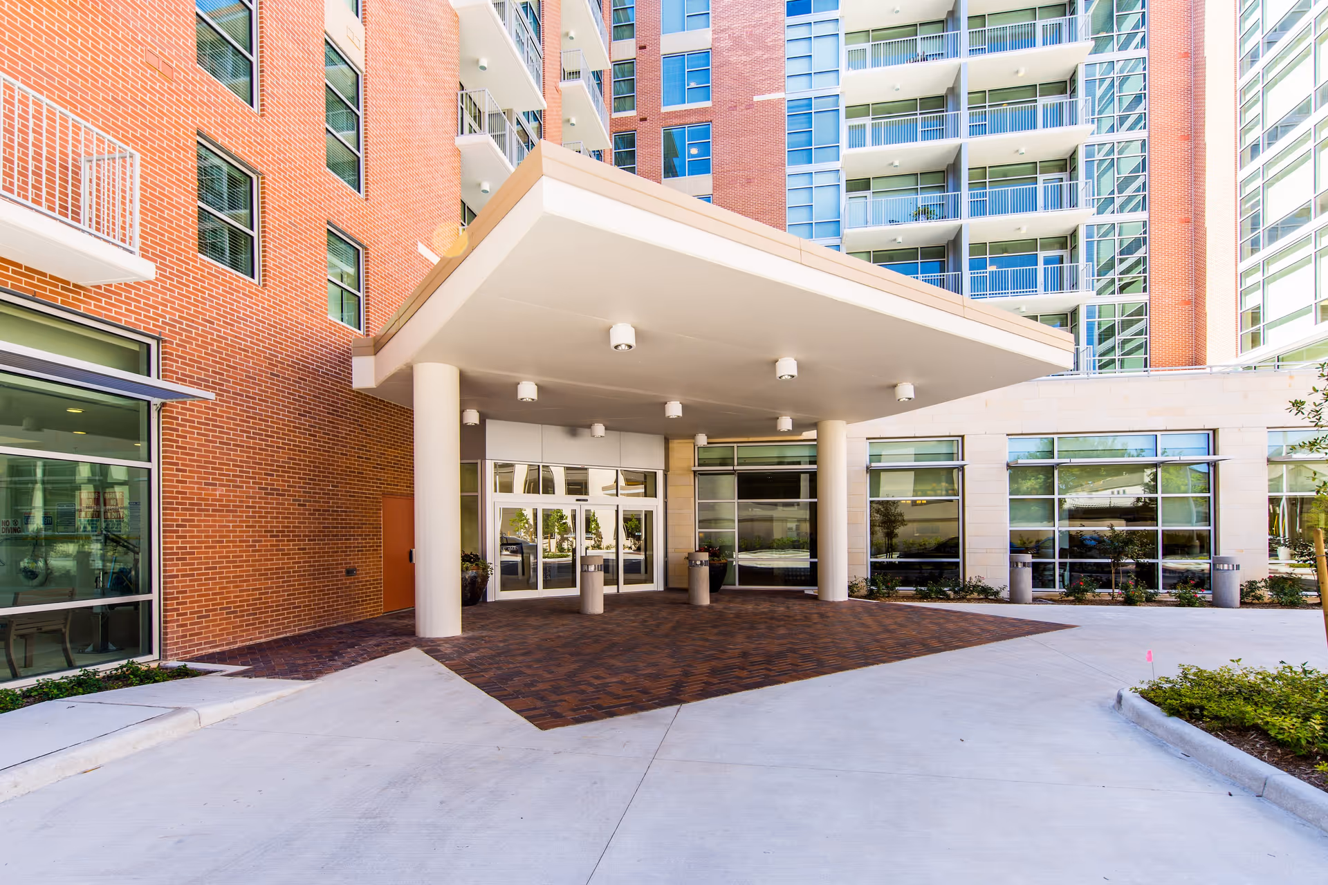Entrance area of a modern multi-story building with a covered drop-off zone supported by white columns. The building features large windows, balconies, and a combination of brick and light-colored exterior walls. There are plants and shrubs along the perimeter near the windows.