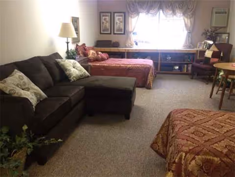 Cozy senior living room with a brown sofa, two beds, a table, and windows.
