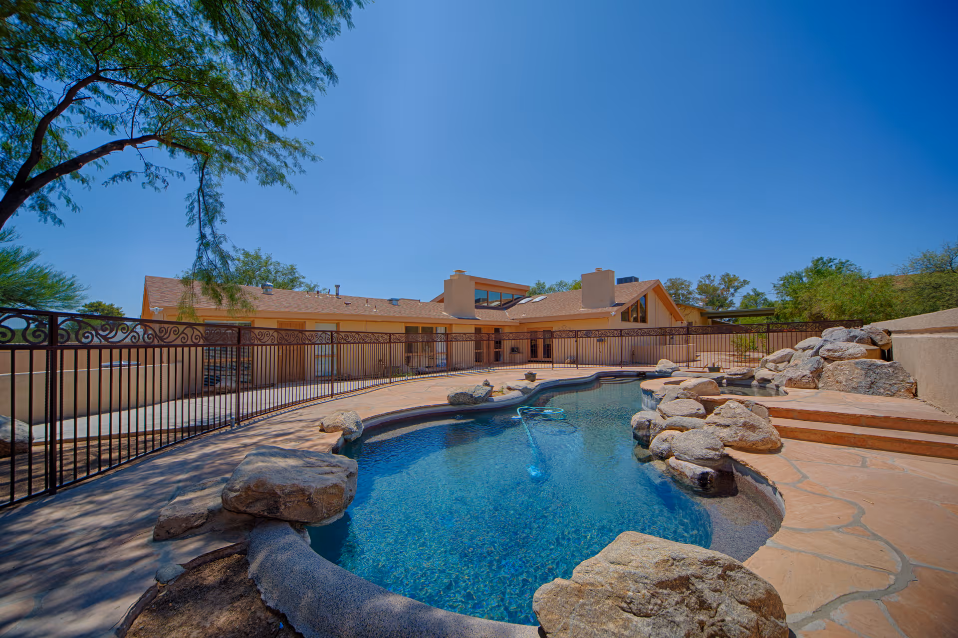 Outdoor swimming pool surrounded by natural rocks and a stone patio, with a beige building and clear blue sky in the background.