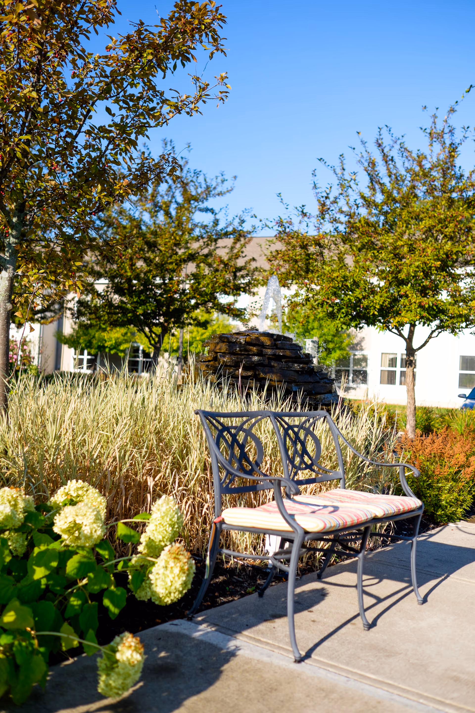 Outdoor garden area with a metal bench featuring a striped cushion, surrounded by green plants and flowers. In the background, there is a stone water fountain and several trees under a clear blue sky. A building is partially visible behind the trees.