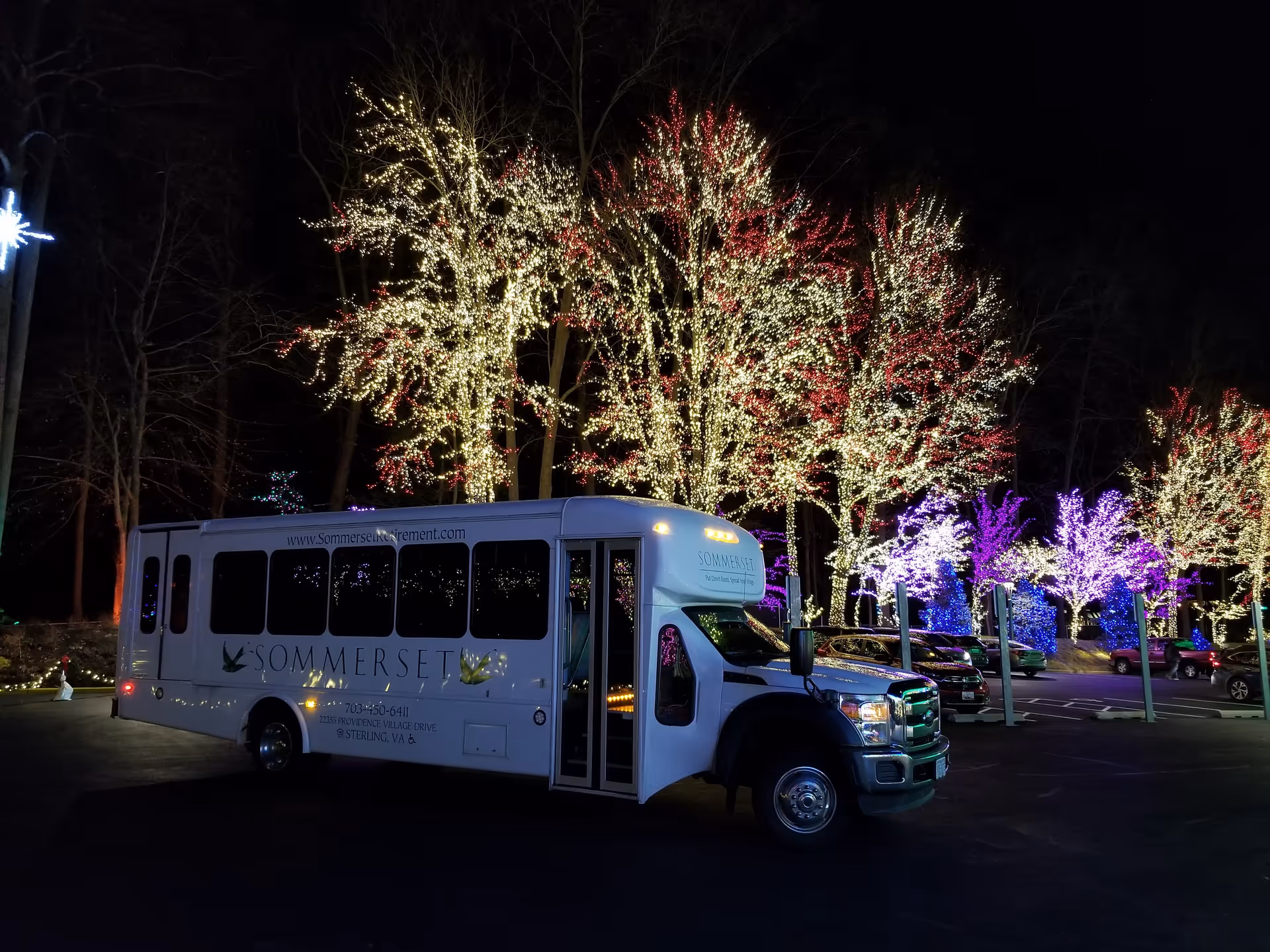 A Sommerset Retirement Community shuttle bus parked at night in a lot with trees wrapped in multicolored holiday lights.