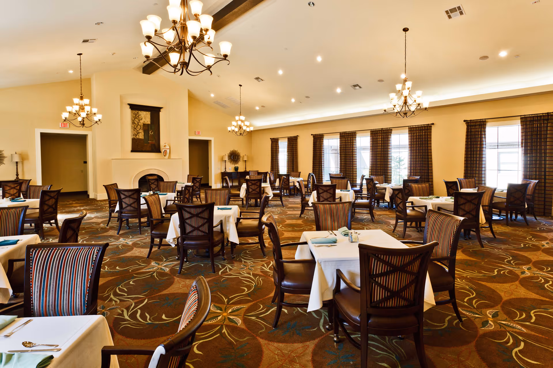 A spacious dining room with multiple tables covered in white tablecloths, each set with napkins and silverware. The room features large windows with patterned curtains, chandeliers hanging from the ceiling, and a decorative fireplace on one wall. The carpet has a floral pattern, and the overall ambiance is warm and inviting.