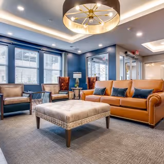 A modern living room with a large tan leather sofa adorned with dark blue pillows, two brown armchairs, and a large tufted ottoman in the center. The room features blue walls, large windows with blinds, a stylish ceiling light fixture, and a gray carpeted floor.