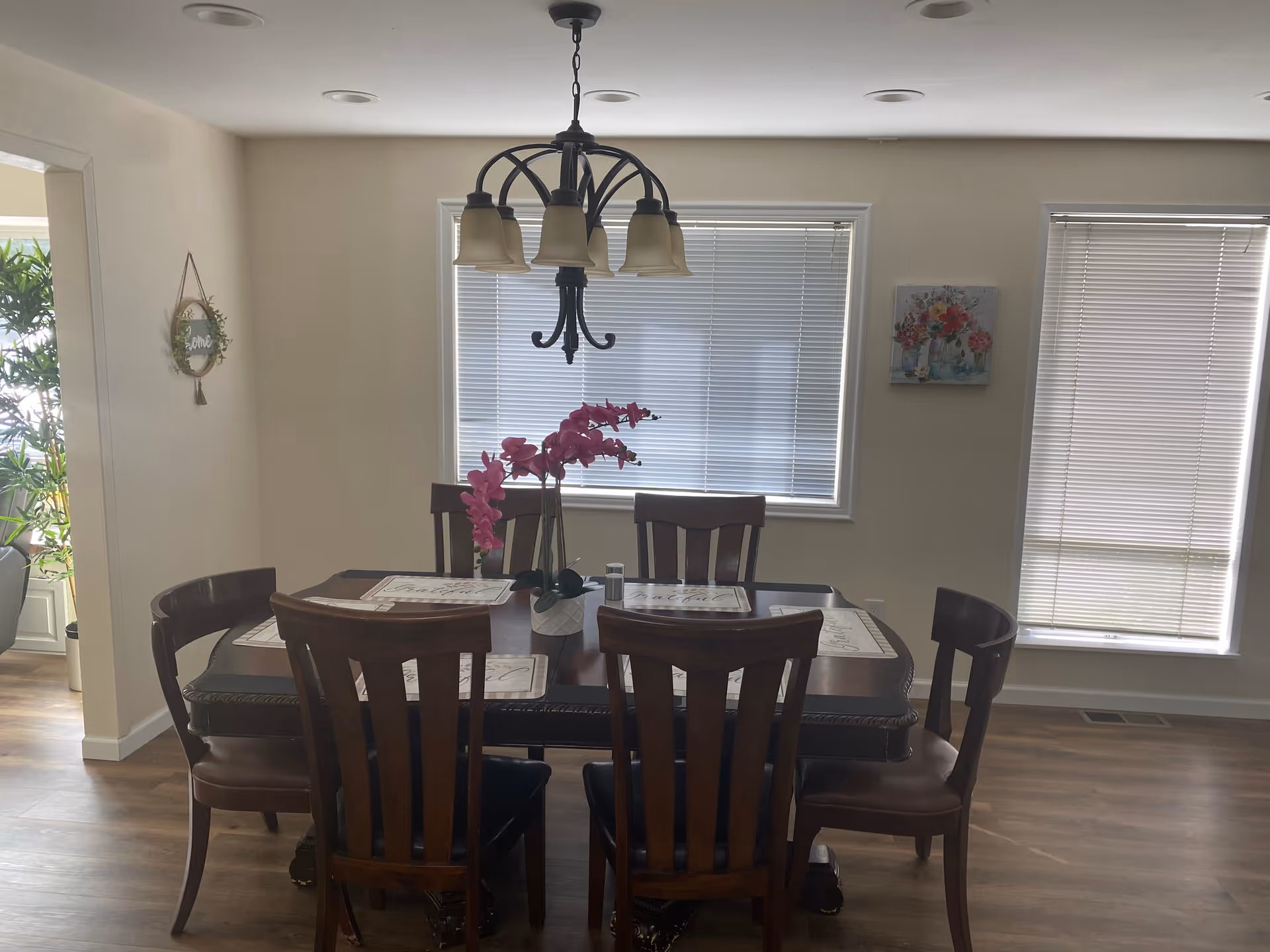 A dining room with a dark wooden table surrounded by six matching chairs. The table has placemats and a vase with pink flowers in the center. There is a chandelier with five lights hanging above the table. Two windows with closed blinds are on the wall behind the table. The room has light-colored walls and wooden flooring. A small wall decoration and a painting with flowers are visible on the walls.