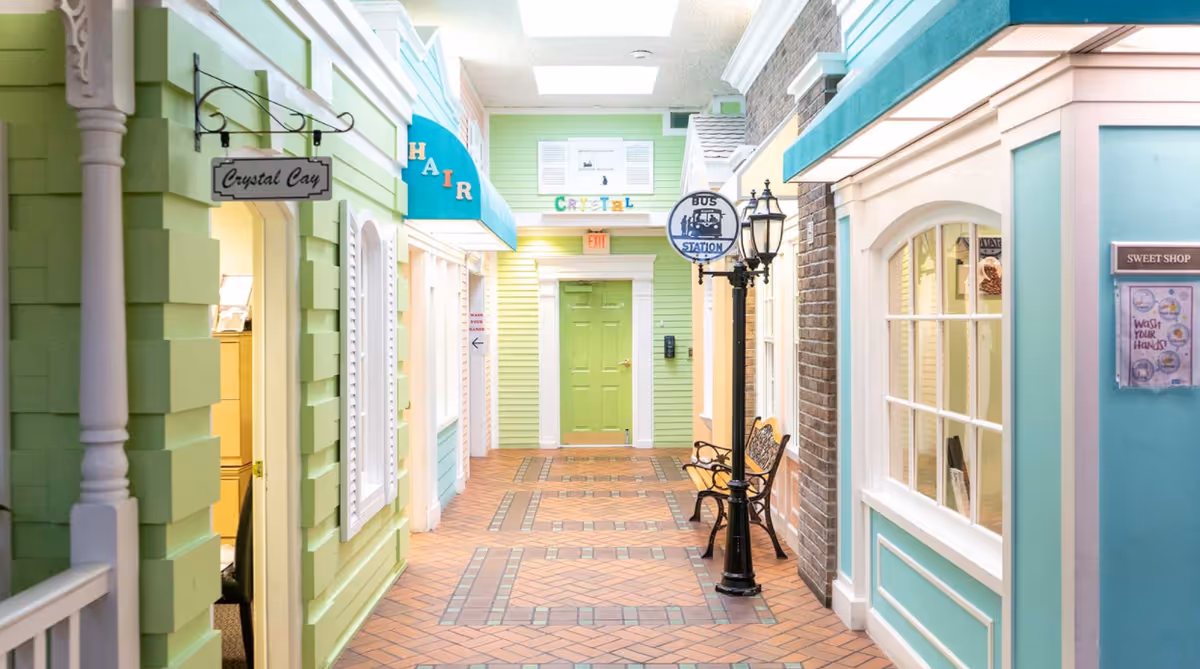 Indoor hallway designed to resemble a quaint village street with pastel-colored storefronts including signs for Crystal Cay, Hair, and Sweet Shop. The hallway features a tiled floor, a green door at the end, a vintage-style street lamp with a bus station sign, and benches along the wall.