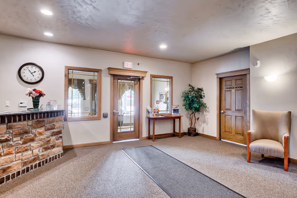 Interior view of a senior living facility lobby area with a stone reception desk on the left, a wall clock above it, a glass door with an exit sign above it in the center, a wooden table with brochures and a potted plant next to it, and a beige armchair near a wooden door labeled 'Family Room' on the right.